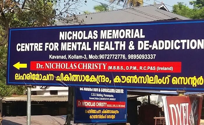 Large blue roadside board reading Nicholas Memorial Centre for Mental Health and De-Addiction, Kavanad, Kollam-3, with phone numbers, Dr. Nicholas Christy's name and a yellow arrow